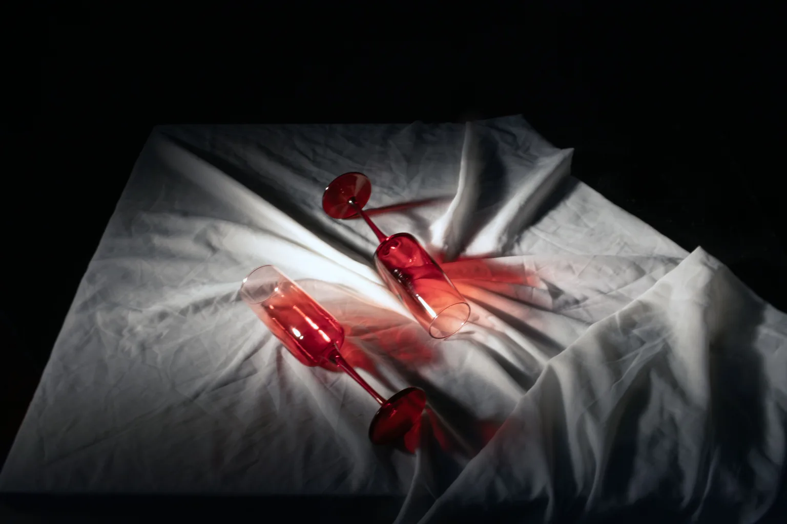 Two tipped wine glasses casting red shadows across a white draped cloth