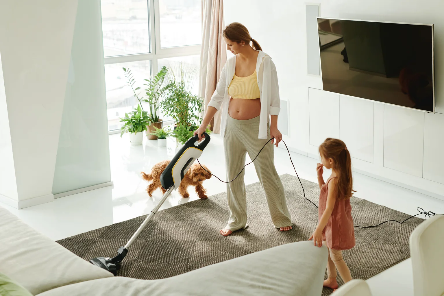 A person vacuuming a living room carpet while a child and dog play nearby
